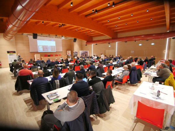 Conferenza con persone sedute a tavoli, schermo proiettato.