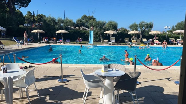 Persone nuotano in piscina, tavoli e sedie in primo piano.