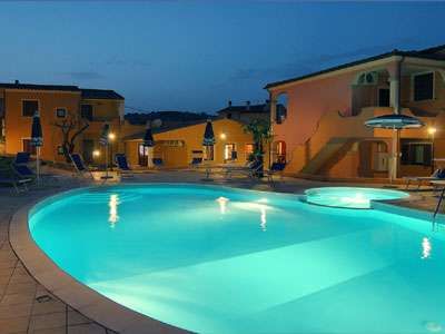 Piscina accesa di sera, con ombrelloni intorno e edifici sullo sfondo.