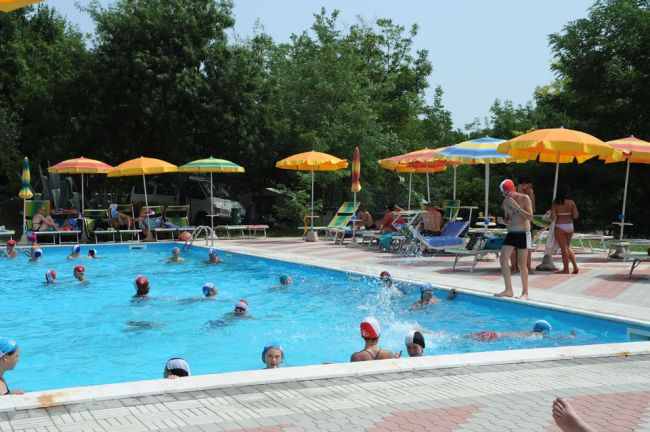 Piscina affollata con persone che nuotano, circondata da ombrelloni gialli e arancioni.