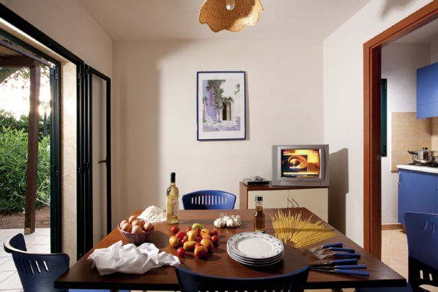 Una cucina con un tavolo apparecchiato con frutta, uova, vino e pasta, e una piccola TV accesa.
