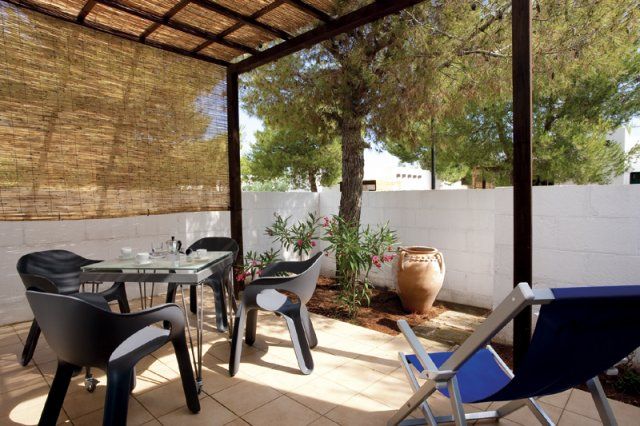Un patio con sedie nere, tavolo di vetro, piante e una sdraio blu sotto una tettoia di canne.