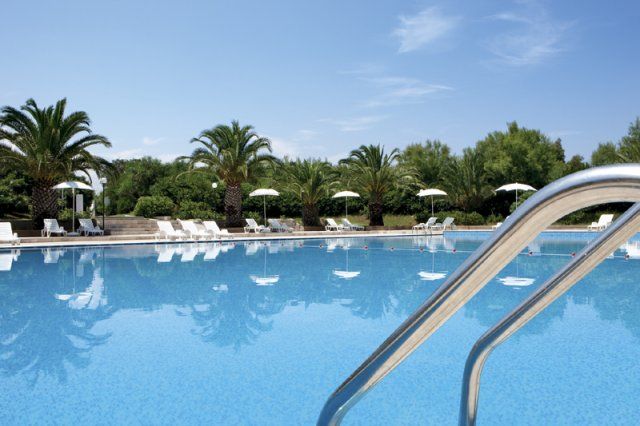 Piscina azzurra con palme e ombrelloni bianchi sullo sfondo, cielo sereno.