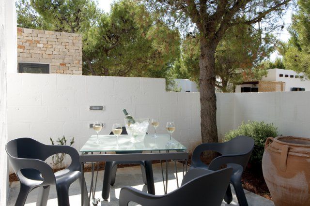 Tavolo con sedie nere e bicchieri di vino su terrazza, circondato da alberi e muro.