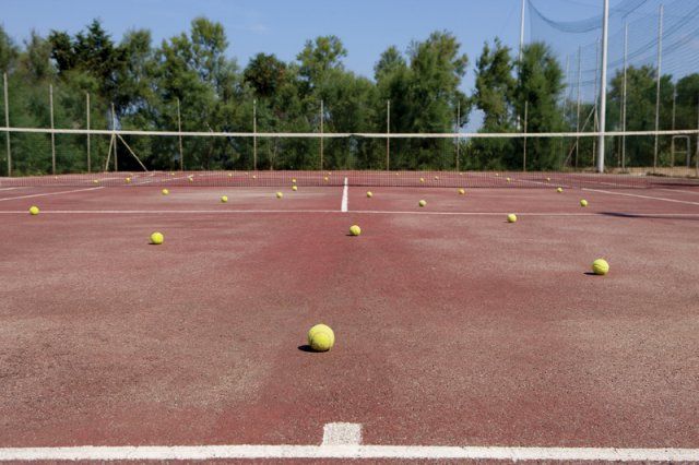 Campo da tennis rosso con palline gialle sparse, circondato da alberi e rete.