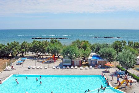 Una piscina vicino alla spiaggia, circondata da alberi e sdraio. Il mare si vede sullo sfondo.