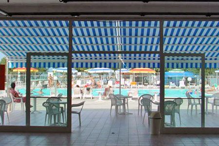 Piscina vista dall'interno, con sedie e ombrelloni colorati all'esterno.