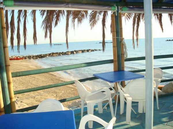 Tavoli e sedie sotto un tetto di paglia, vista sul mare con scogli.