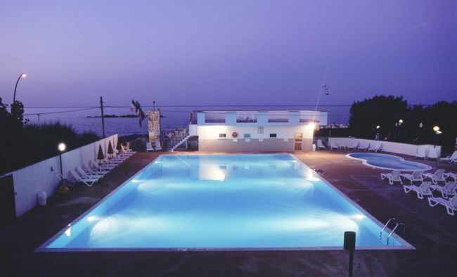 Piscina illuminata al tramonto, sdraio intorno e mare sullo sfondo.