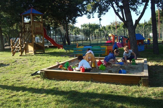 Bambini che giocano in una sabbiera in un parco giochi con alberi e attrezzature colorate.