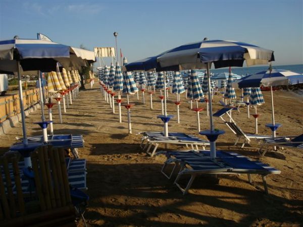 Spiaggia con ombrelloni blu e bianchi allineati su sabbia, lettini da sole vuoti.