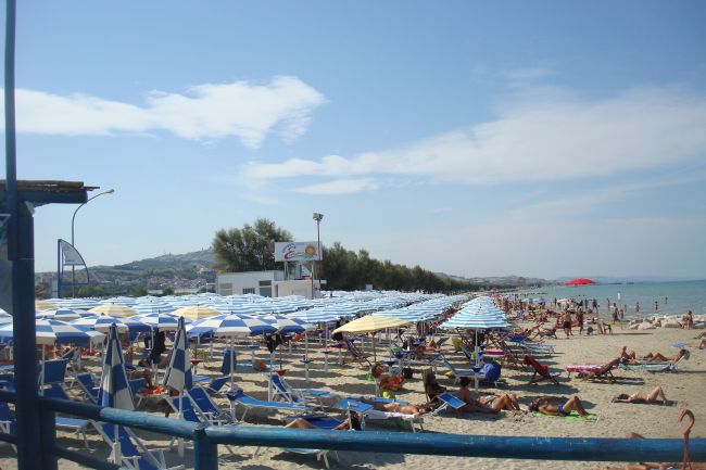 Spiaggia affollata con persone su lettini sotto ombrelloni blu e bianchi.
