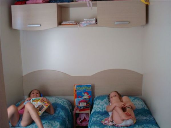 Due bambini su letti singoli leggono in una stanza con pareti chiare e mobili sopraelevati.