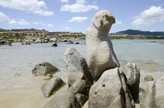 Rocce scolpite emergono dal mare limpido con case sullo sfondo.
