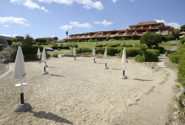 Spiaggia sabbiosa con ombrelloni chiusi, villette sullo sfondo.
