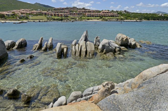 Scogli affioranti in mare cristallino, complesso residenziale sullo sfondo.
