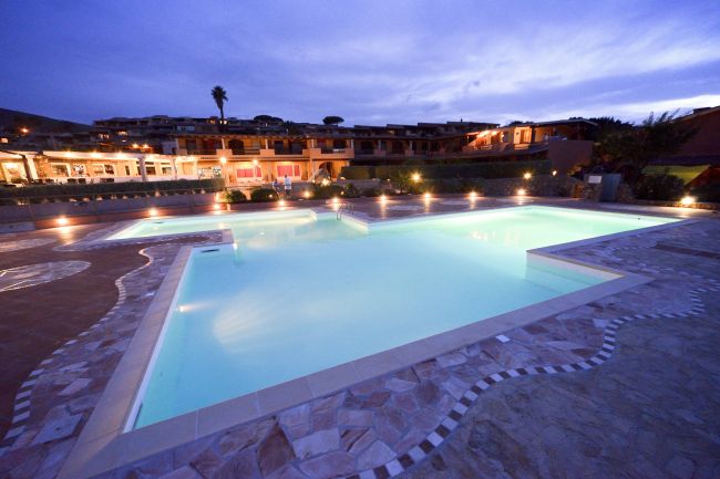 Piscina illuminata di sera con edifici sullo sfondo.