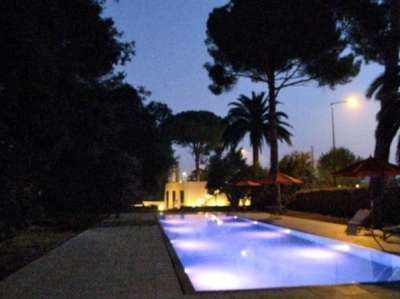 Piscina illuminata di notte con luci blu, circondata da alberi e ombrelloni.