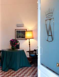 Una sala con tavolo coperto da tovaglia verde, fiori e lampada. Una porta con stemma è aperta.