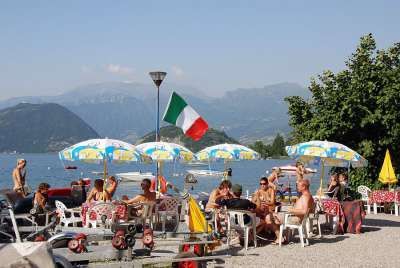 Persone sedute a un bar all'aperto vicino a un lago, con ombrelloni e bandiera italiana.