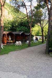 Sentiero in ghiaia con casette in legno e sedie bianche da giardino tra alberi.