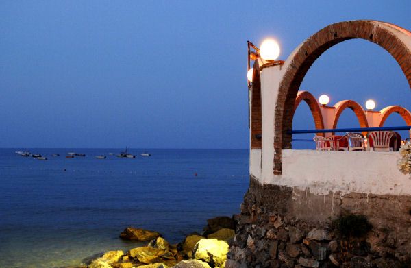Terrazza con archi e sedie sul mare al tramonto, vista di barche e acqua.