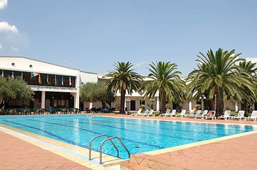 Piscina all'aperto con lettini e palme, edificio sullo sfondo.