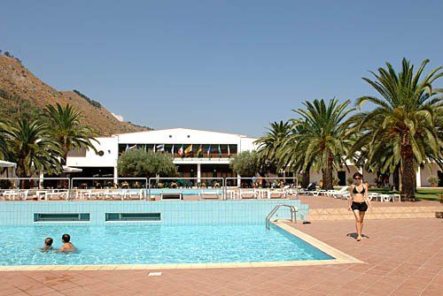 Piscina con persone che nuotano, palme sullo sfondo e un edificio bianco.