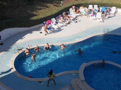 Gruppo di persone nuota in una piscina, altre prendono il sole su sdraio.
