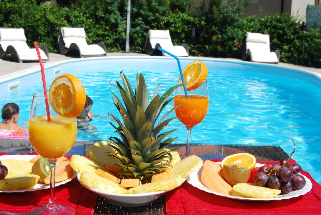 Due cocktail con fette d'arancia e piatti di frutta davanti a una piscina.