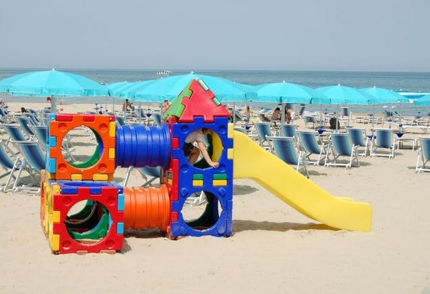 Gioco colorato per bambini su una spiaggia con sedie a sdraio blu e ombrelloni sullo sfondo.