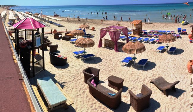 Spiaggia con lettini in vimini, ombrelloni di paglia e una struttura rosa vicino al mare.