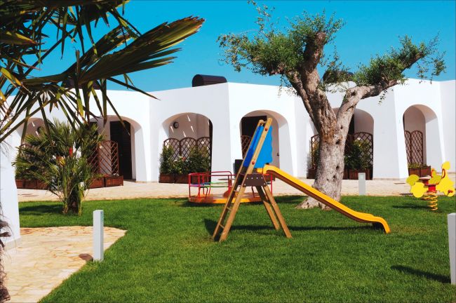 Area giochi su erba con scivolo e giostra, più edifici bianchi sullo sfondo.