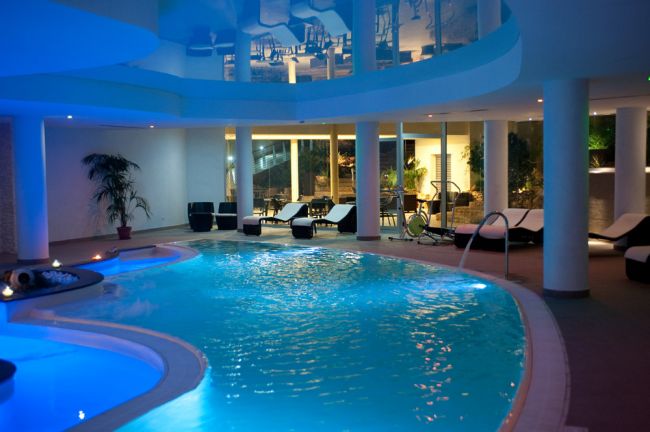 Piscina interna illuminata, circondata da lettini e piante, con struttura moderna a colonne.