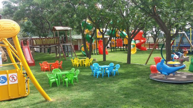 Area giochi con tavolini colorati, scivoli e giostre tra gli alberi.