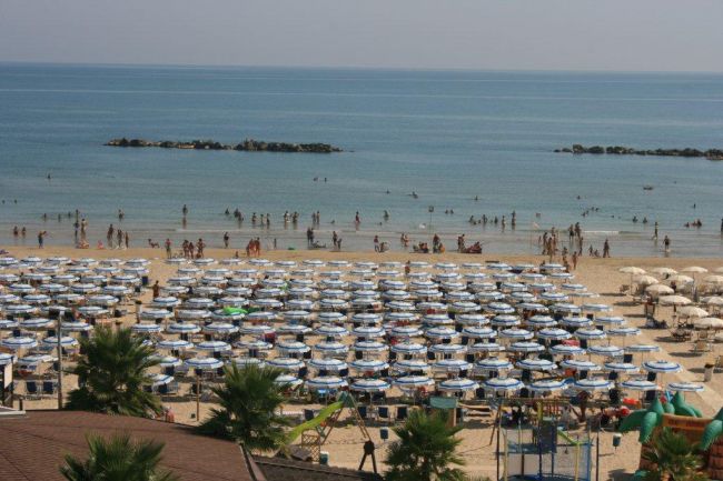 Spiaggia affollata con ombrelloni allineati, mare calmo sullo sfondo.