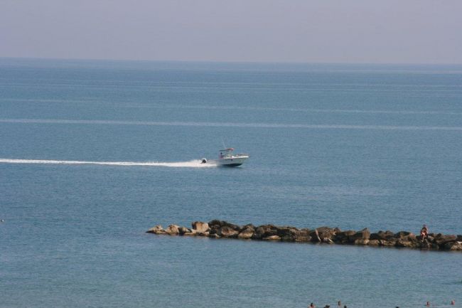 Barca che naviga veloce su un mare calmo vicino a una barriera di rocce, cielo chiaro all'orizzonte.