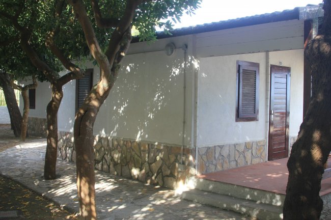 Bungalow bianco con porta in legno, ombreggiato da alberi.