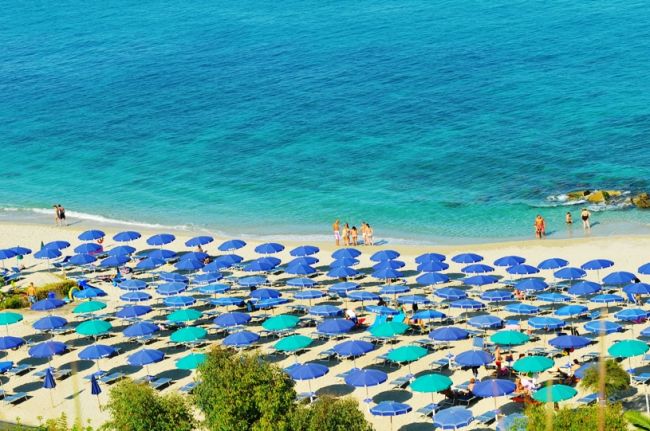 Spiaggia sabbiosa con numerosi ombrelloni blu e mare turchese.