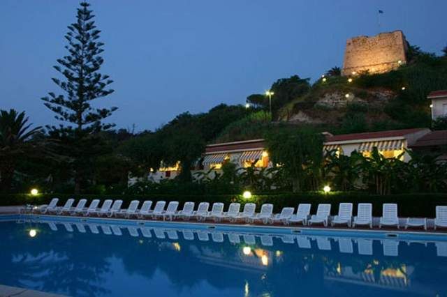 Piscina di notte con sedie a sdraio bianche e una collina con edificio illuminato sullo sfondo.