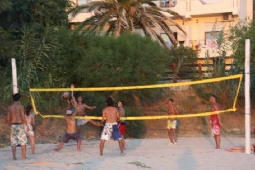 Persone giocano a beach volley su una spiaggia, circondate da vegetazione e palme.