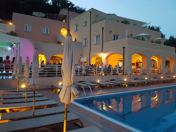 Piscina illuminata di sera, con lettini e edificio sullo sfondo.