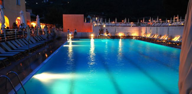 Piscina illuminata di sera con lettini e ombrelloni.