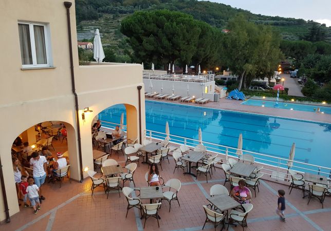 Piscina all'aperto con tavolini, alberi sullo sfondo.