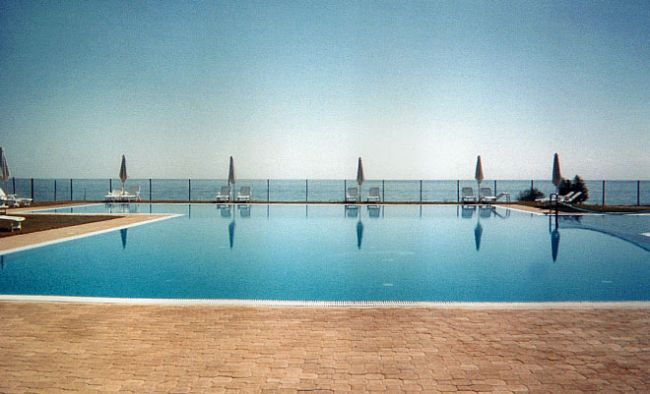 Piscina con vista sul mare, ombrelloni e sdraio sullo sfondo.
