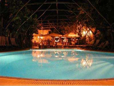 Piscina illuminata di notte, con sedie a sdraio attorno e un bar sullo sfondo.
