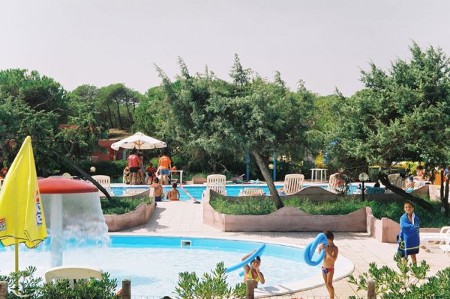 Piscina con bambini che giocano e un fungo d'acqua, circondata da alberi e sdraio.