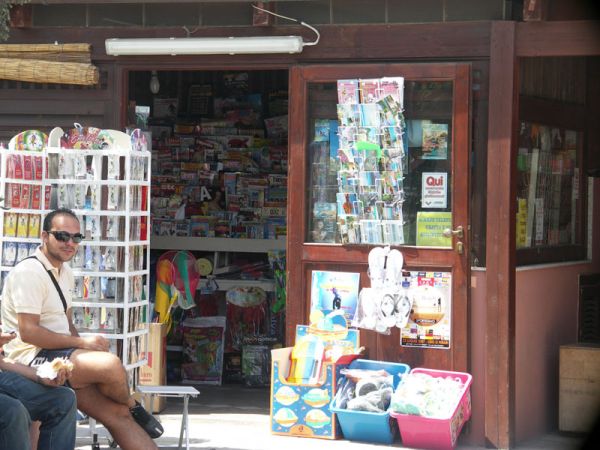 Edicola con cartoline e giornali esposti, uomo seduto davanti al negozio.