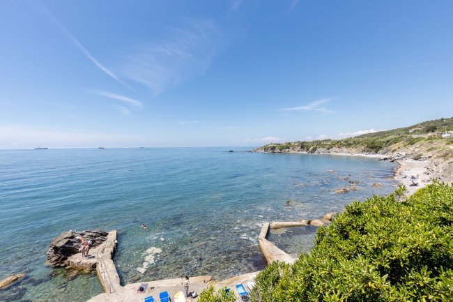 Spiaggia rocciosa con mare calmo e cielo sereno.