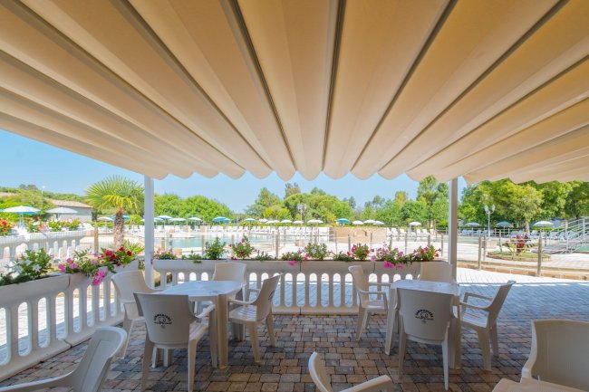 Tavoli e sedie bianche sotto una tenda, con vista su una piscina e giardino fiorito.